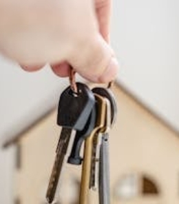 Close-up of a hand holding keys with a miniature wooden house in the background, symbolizing real estate investment.